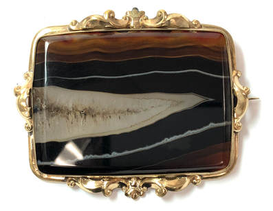 This antique, Victorian era brooch circa the late 1800s, features a large banded agate set at the center of a gold-filled frame with repoussé detailing